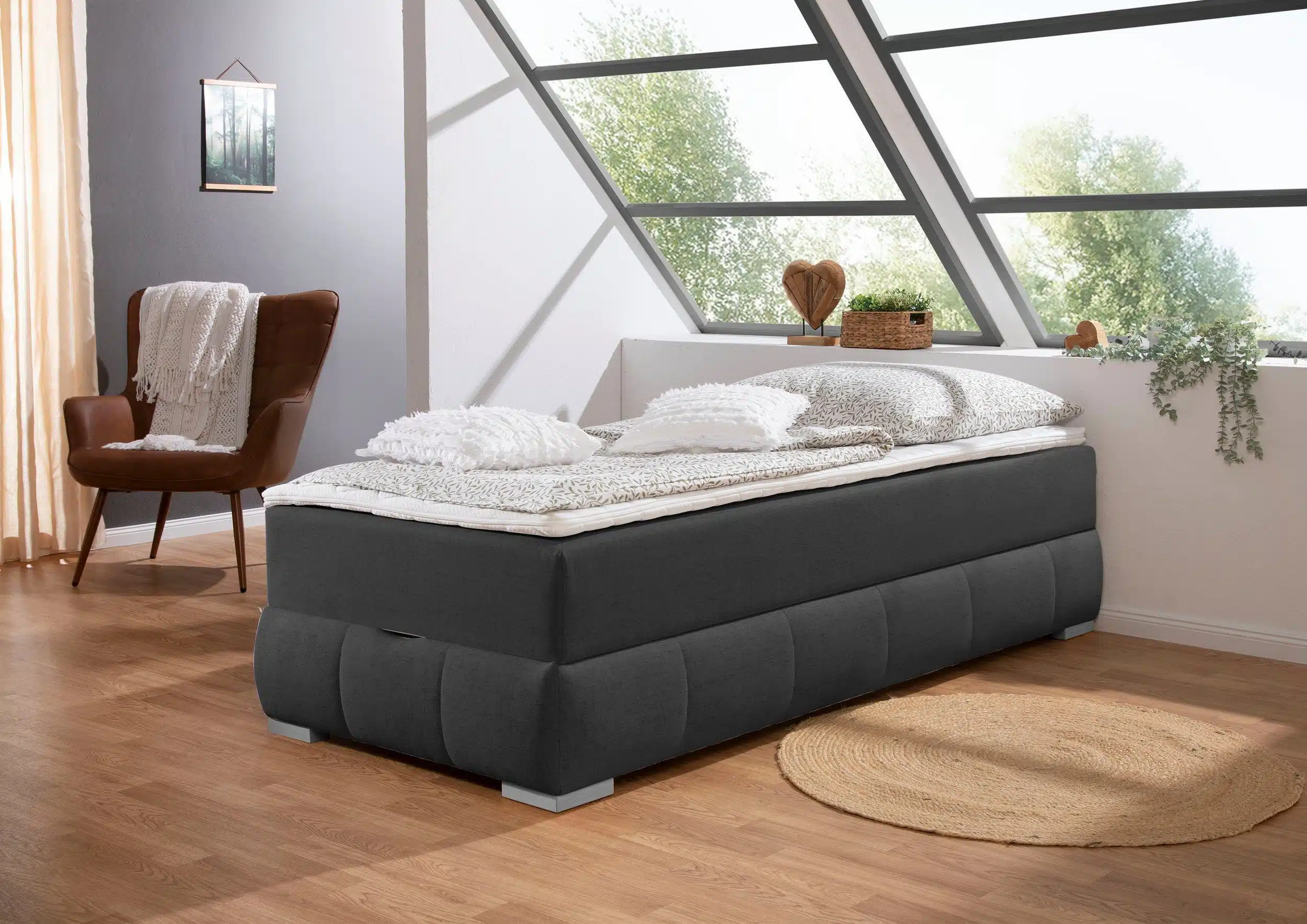 Guido Maria Kretschmer Boxbett in einem stilvollen Schlafzimmer, mit einer gemütlichen Bettwäsche aus hellen Tönen. Ein brauner Sessel und ein runder Teppich aus Jute ergänzen die moderne Einrichtung, während große Fenster für viel Tageslicht sorgen.