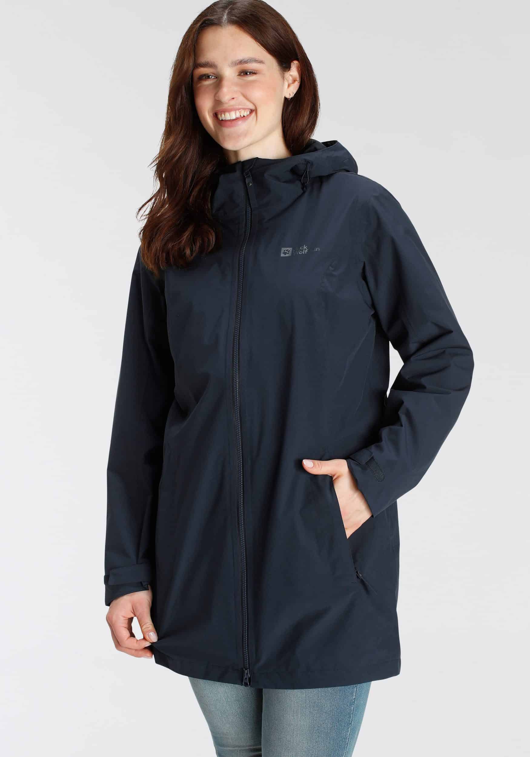 Eine Frau trägt einen blauen Jack Wolfskin Funktionsparka mit Kapuze und Reißverschluss. Sie lächelt freundlich und steht vor einem neutralen Hintergrund.