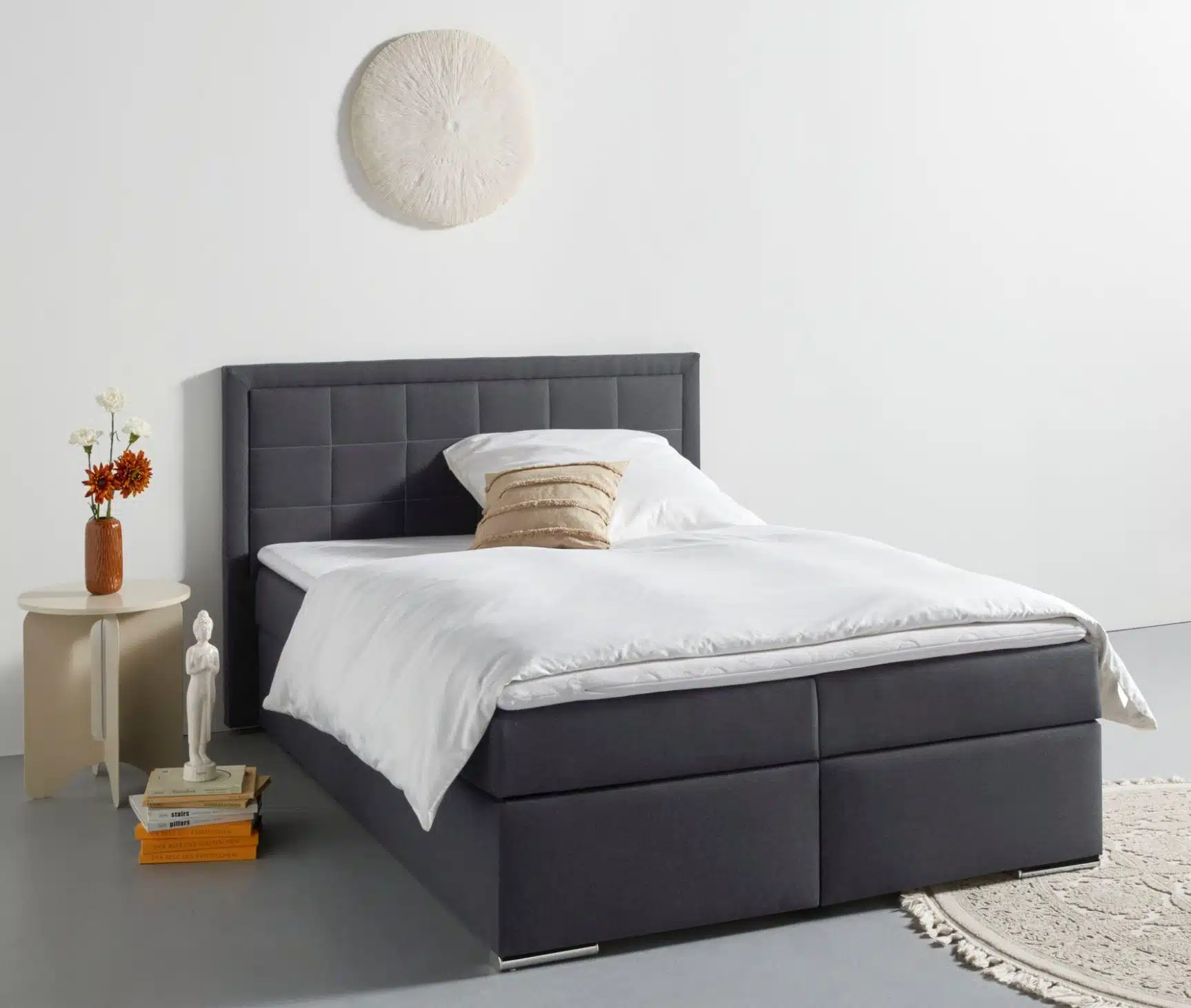 Modern eingerichtetes Schlafzimmer mit einem dunkelgrauen Boxspringbett, weißer Bettwäsche, Beistelltisch mit Deko und rundem Teppich auf grauem Boden.