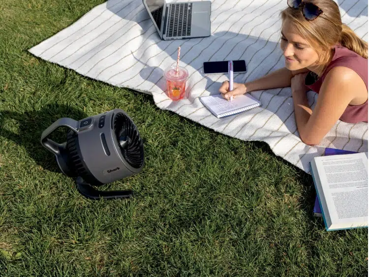 Frau liegt auf ihrer Decke im Park, macht Notizen und genießt den Shark Flex Breeze HydroGo Ventilator mit Sprühnebel.