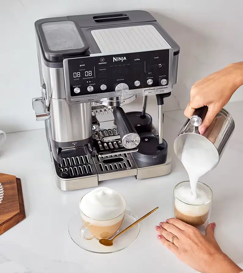 Zu sehen ist die Ninja Luxe Café Pro Kaffeemaschine - ES701EU, dazu wird aus dem Milchaufschümer Milch in den Kaffee gekippt. Daneben steht bereits schon ein fertiger Latte Macchiato.