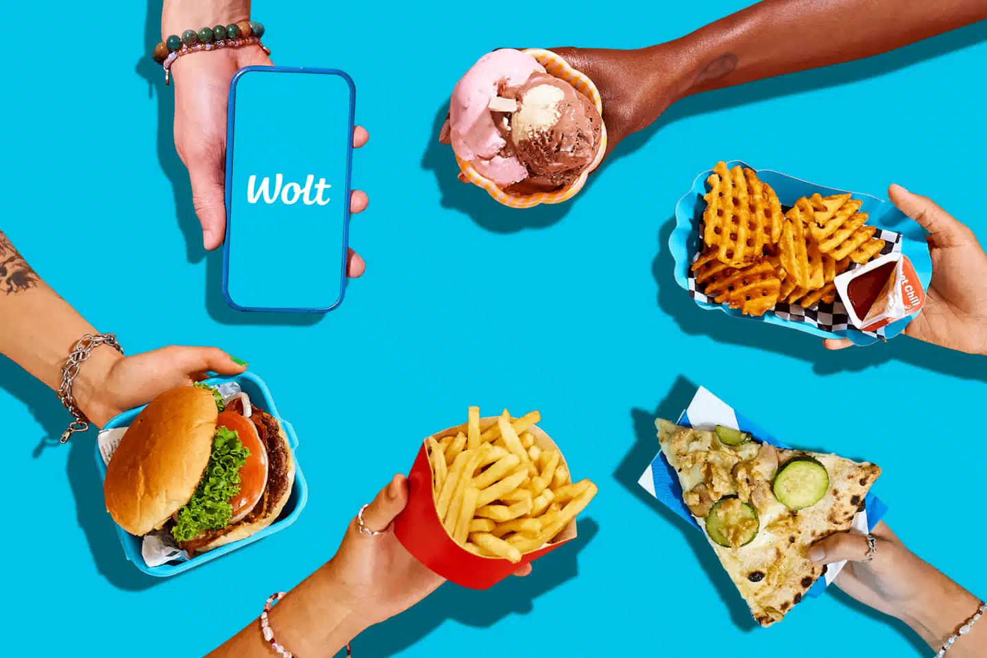 Verschiedene Gerichte bei Wolt in Händen, eine Hand hält ein Handy mit Wolt Schriftzug auf dem Display
