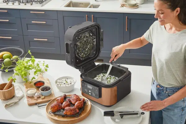 Ninja Speedi Multikocher auf einer Anrichte in der Küche neben frittierten Hähnchenschenkeln und weiteren Gerichten - eine Frau rührt gerade ein Gericht im Multikocher um